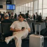 Man sitting on his phone, waiting at an airport terminal gate with a very crowded seating section around him.